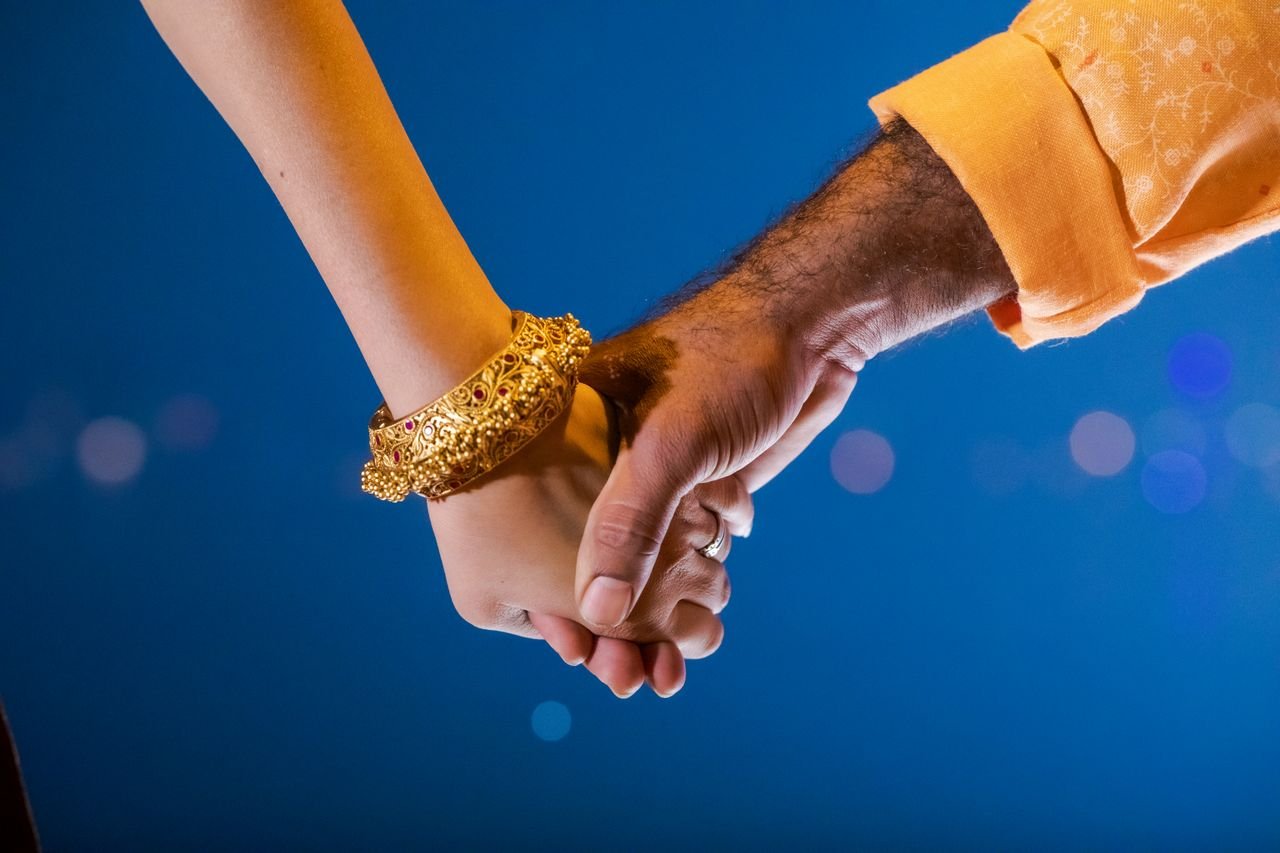 Close-up of a couple holding hands, featuring traditional Indian jewelry against a vibrant blue background.
